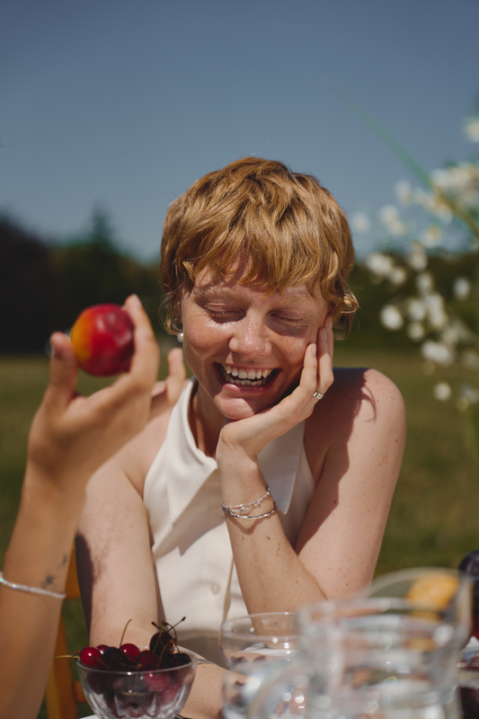 Усміхнена дівчина з короткою рудою стрижкою у білому топі насолоджується літнім пікніком, срібні прикраси на руці